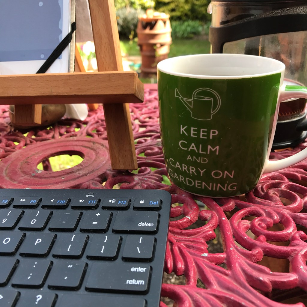 A coffee cup on a red bistro table in the garden, with keyboard and tablet ready for a gardening ways blog article